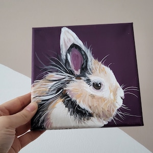 May include: A painting of a fluffy white rabbit with black and brown markings on a purple background. The rabbit has a pink nose and brown eyes.