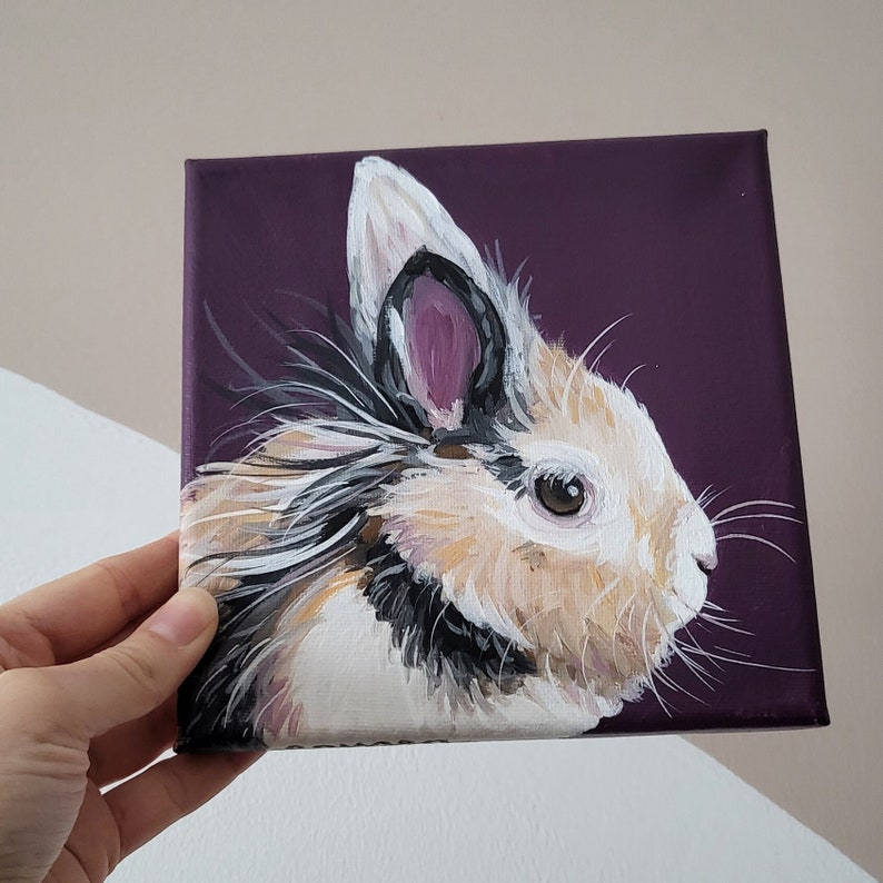 May include: A painting of a fluffy white rabbit with black and brown markings on a purple background. The rabbit has a pink nose and brown eyes.