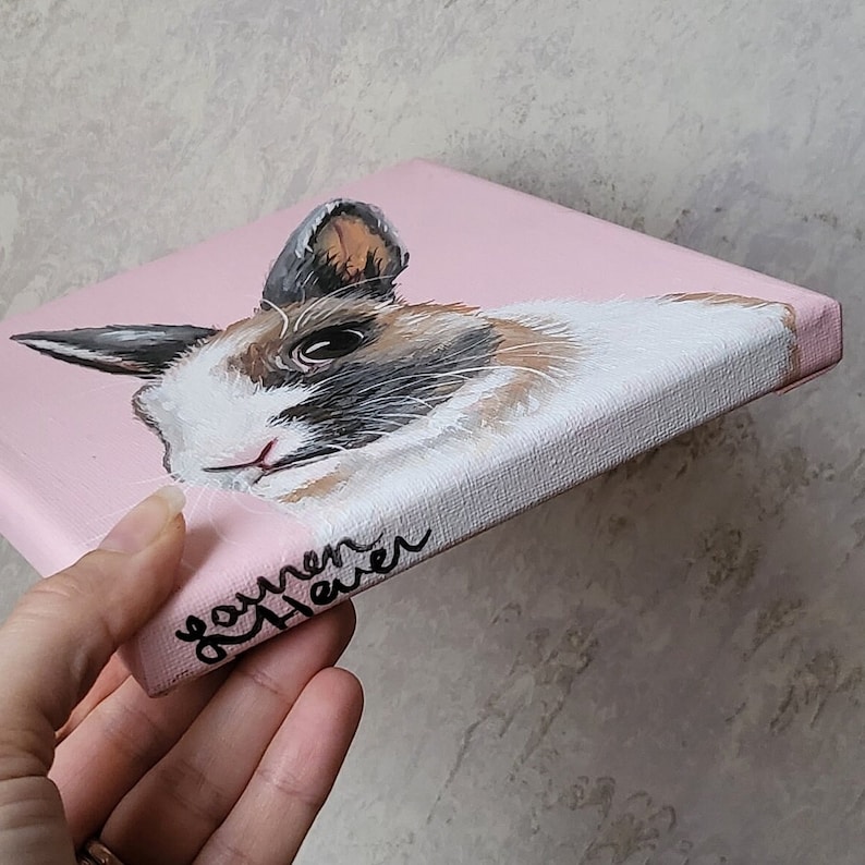 May include: A hand-painted canvas featuring a close-up portrait of a brown and white rabbit with black markings. The rabbit has a pink nose and large, dark eyes. The canvas is painted on a pink background and has the name "Lauren Haver" written on the side in black.