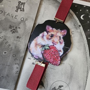 May include: A bookmark featuring a hamster holding a strawberry. The hamster is brown and white, set against a black background. The bookmark has a red elastic ribbon with gold accents. The bookmark is on a map.