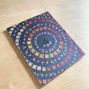 May include: A circular design made of pressed dried flowers and leaves on a dark blue background. The design features a variety of colors, including red, orange, yellow, and green.