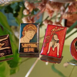May include: Four vintage enamel pins with various designs. One pin features a reclining figure, another has a portrait with the text "Staying Alive", a third pin displays a woman with the text "Flashdance", and the last pin has a silhouette with the text "Michael Jackson".
