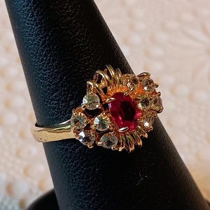 May include: A gold ring featuring a central, oval-cut, deep red gemstone surrounded by smaller, clear gemstones. The ring's setting has a floral or sunburst design, with intricate gold detailing. The ring is displayed on a black ring holder.