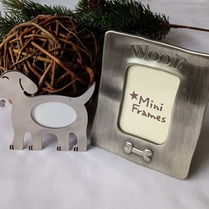May include: Silver metal picture frame shaped like a dog with a bone in its mouth. The frame has a small oval opening for a photo. The frame is next to a silver metal square picture frame with the words "Woof Mini Frames" on it.
