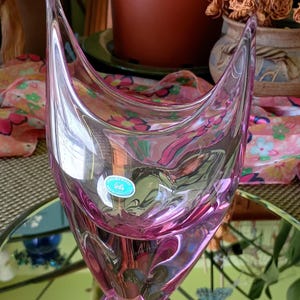 May include: A pink glass vase with a unique, curved design. The vase has a wide opening and a rounded base. The glass is transparent, allowing light to pass through and reflect off the curves. A small, round sticker is visible on the front of the vase.