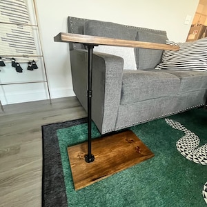 May include: A C-shaped side table featuring a rectangular dark wood top and a matching rectangular wood base connected by a single vertical black metal pipe support. The table is designed to slide under furniture for convenient surface access.