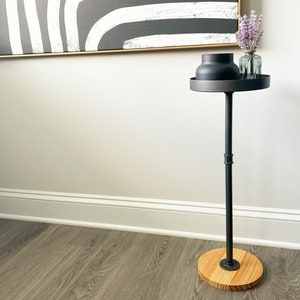 May include: A black metal side table with a wooden top. The table has a single round base and a single round top. A black vase and a glass vase with dried flowers are on the table.