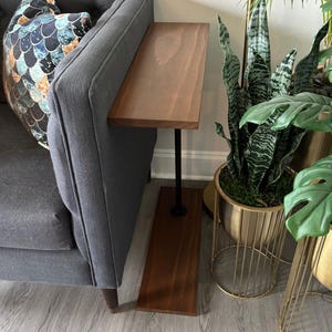 May include: A narrow C-shaped side table with a rectangular wooden top and base connected by a single black metal pole. The table is positioned beside a dark gray sofa featuring a patterned throw pillow with a blue and gold scale design.