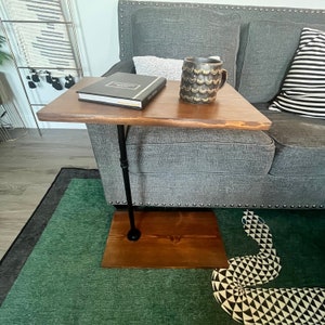 May include: A C-shaped side table with a rectangular dark brown wooden top and base connected by a single black metal pipe support. A black book with white text and a dark textured mug with gold scalloped patterns sit on the wooden surface.