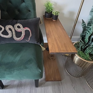 May include: A dark green velvet chair with a decorative pillow featuring a snake design. A wooden side table with two small potted plants sits next to the chair. The table is stained a warm brown color.