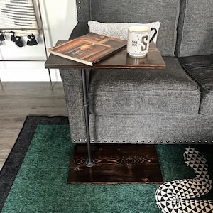 May include: A C-shaped side table featuring a dark brown stained wooden top and base connected by a black metal pipe support. A magazine and a white ceramic mug with a black letter S are placed on the table surface next to a gray upholstered sofa.