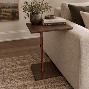 May include: A C-shaped side table featuring a dark brown wooden top and base connected by a single vertical copper-toned metal pipe. The table is positioned beside a sofa, with a dark ceramic pot containing a green plant and two stacked books.