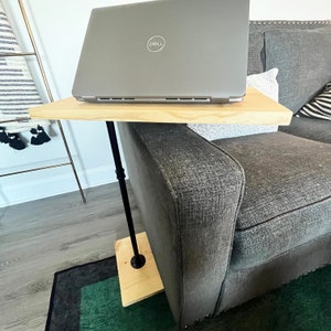 May include: A C-shaped side table featuring a light-toned wooden top and base connected by a slender black metal pole. The table is positioned alongside a sofa, providing a surface for a silver Dell laptop that is resting on the wooden tabletop.