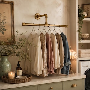 May include: A wall-mounted brass-toned metal pipe clothing rack featuring a horizontal bar with six metal hangers. Hanging from the rack are various long-sleeved shirts in shades of white, floral print, dusty rose, and a blue denim jacket.