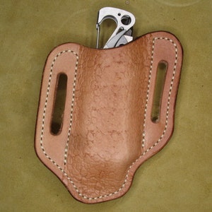 Könnte beinhalten: Eine braune Ledertasche für ein Taschenmesser mit einem genähten Design und einem Metallclip.