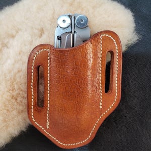 Peut inclure: Un étui en cuir marron pour multi-outils avec des détails de couture blanche. L'étui a deux passants de ceinture et est conçu pour contenir un multi-outils.