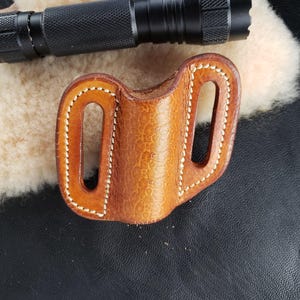 Puede incluir: Una funda de linterna de cuero marrón con costuras blancas. La funda tiene dos trabillas para el cinturón y una sola ranura para una linterna.