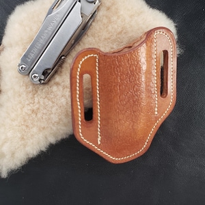 May include: A brown leather multi-tool sheath with white stitching. The sheath has two belt loops and a textured surface.