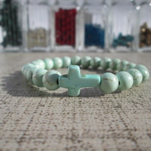 Peut inclure: Un bracelet de perles vert clair avec un charm en forme de croix. Le bracelet est composé de perles rondes et d'une croix, le tout dans une couleur vert clair assortie. Le fond montre des bocaux en verre avec des contenus colorés.