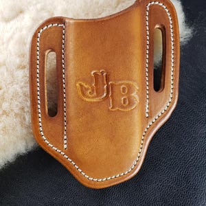 Pode incluir: Uma coldre de couro castanho com costura branca e as iniciais "JB" em relevo. O coldre tem duas ranhuras e está colocado sobre um fundo texturizado. O couro tem um acabamento liso e polido.