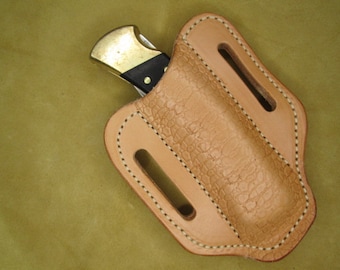 May include: A light brown leather knife sheath with a stitched edge and two belt loops. The sheath is designed to hold a folding knife with a brass handle.