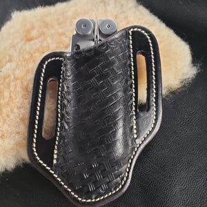 May include: A black leather multi-tool holster with a patterned design and a white stitching detail. The holster has a belt loop and a pocket for a multi-tool.