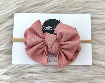May include: A dusty rose ribbed fabric bow headband. The bow is centered on a thin, neutral-coloured elastic band. The bow is knotted in the centre. The product is displayed on a white card with the text "Stella J & Jane Headbands".