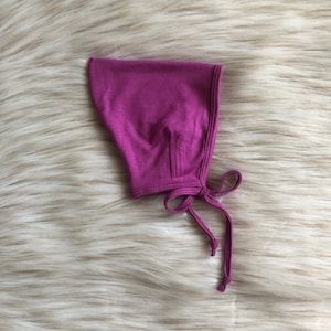 Puede incluir: Un gorro de bebé magenta hecho de tela suave. El gorro tiene una forma redondeada con un cierre de lazo debajo de la barbilla. El fondo es una superficie esponjosa y de color claro.