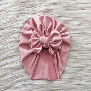 Op de afbeelding: Een stoffige roze turban met een grote strik bovenop. De turban is gemaakt van een zachte, rekbare stof.