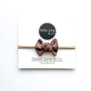 Puede incluir: Una diadema anudada marrón con una banda elástica nude. La diadema está sobre un fondo blanco con el texto "Stella Jane & Co. Headband".