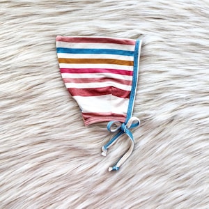 May include: A striped baby bonnet with a blue and white tie. The bonnet is made of a soft, stretchy fabric and has a pink, brown, and white striped pattern.