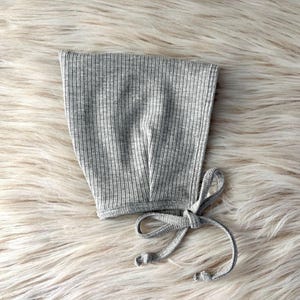 May include: A gray ribbed knit baby bonnet with a tie closure. The bonnet is made of a soft, stretchy fabric and is designed to fit a baby's head. The bonnet is shown on a fluffy, white surface.