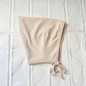 May include: A beige ribbed knit bonnet with a tie closure. The bonnet is made of a soft, neutral-toned fabric and is designed to cover the head and neck. The background is a white brick wall.