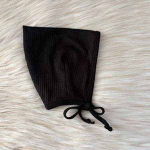 May include: A black ribbed knit baby bonnet with a tie fastening. The bonnet is made from a soft, stretchy fabric and is designed to fit comfortably on a baby's head. The bonnet is a solid black colour.