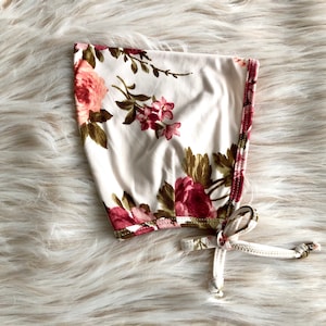 Puede incluir: Un gorro blanco floral con un estampado floral rosa y verde. El gorro tiene un cierre de lazo.