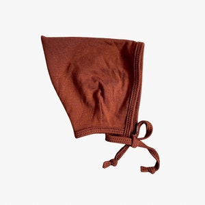 Puede incluir: Un gorro de bebé marrón con un lazo. El gorro está hecho de una tela suave y elástica.