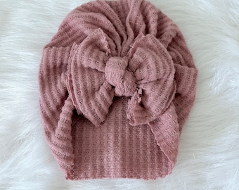 May include: A mauve knitted turban with a large bow on top. The turban is made of a soft, textured fabric.