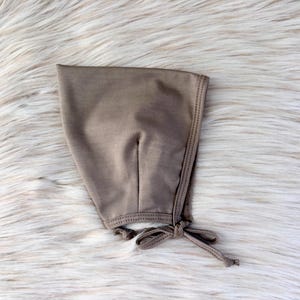 May include: A taupe-colored baby bonnet made of soft fabric. The bonnet has a rounded shape with a seam down the center and a delicate tie closure at the chin. The edges are finished with a contrasting stitch.