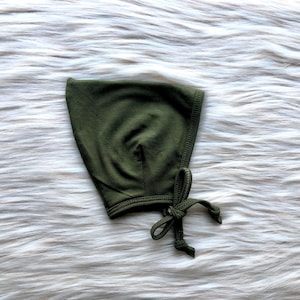 Può includere: Un cappellino da neonato verde oliva realizzato in tessuto morbido. Il cappellino ha una forma arrotondata con una chiusura a fiocco sotto il mento. Il tessuto sembra essere un materiale a maglia e il cappellino è posto su uno sfondo bianco e soffice.