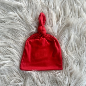 May include: A red knotted baby hat made of soft fabric. The hat is on a white fluffy surface.