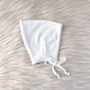 May include: A white, ribbed knit baby bonnet with a tie closure. The bonnet is made of a soft, textured fabric and is designed to cover the head and neck. The background is a fluffy, light-colored surface.