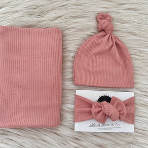 Op de afbeelding: Een set van drie babyaccessoires in een stoffige roze kleur. De set bevat een geribbelde gebreide muts, een bijpassend hoofdband en een swaddle-deken.