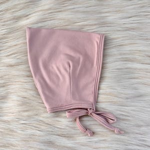 May include: A dusty rose-colored baby bonnet with a tie closure. The bonnet is made of soft fabric and features decorative stitching along the edges. The bonnet is laying on a fluffy, white fur-like surface.