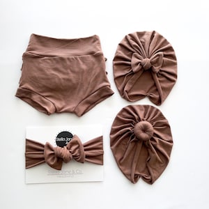 May include: A set of three brown baby accessories: a pair of ruffled shorts, a headband with a bow, and a turban with a bun. The headband is labelled "Stella Jane & Co. Headband".
