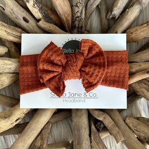 Könnte beinhalten: Ein rostiges Stirnband mit einer großen, geknoteten Schleife. Das Stirnband ist aus einem weichen, strukturierten Stoff gefertigt. Der Text "Stella Jane & Co. Headband" ist auf einer weißen Karte hinter dem Stirnband gedruckt.