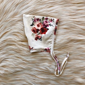 May include: A white bonnet with a floral print featuring red, pink, and green flowers. The bonnet has a tie that can be tied under the chin.