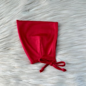 May include: A vibrant red baby bonnet with a tie closure. The bonnet is made of a soft, smooth fabric and is designed to cover the head and neck. The background is a fluffy, white surface.