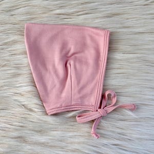 Pode incluir: Um gorro de bebê rosa claro feito de tecido macio. O gorro tem um formato arredondado com uma borda costurada e um fecho de laço. O gorro é projetado para proteger a cabeça do bebê.