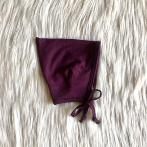 Può includere: Un cappuccio da bambino bordeaux scuro con chiusura a fiocco. Il cappuccio è realizzato in un tessuto morbido ed è posto su uno sfondo bianco e soffice.