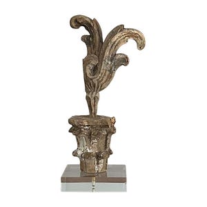 May include: A weathered wooden finial featuring a carved acanthus leaf design atop a fluted base, mounted on a clear square acrylic stand. The wood displays a distressed, muted brown and grey finish with visible signs of age and texture.
